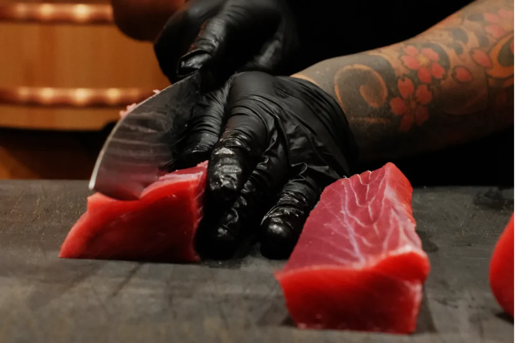 A person wearing black gloves and a floral tattoo is slicing fresh tuna on a dark cutting board, highlighting careful preparation and precision.