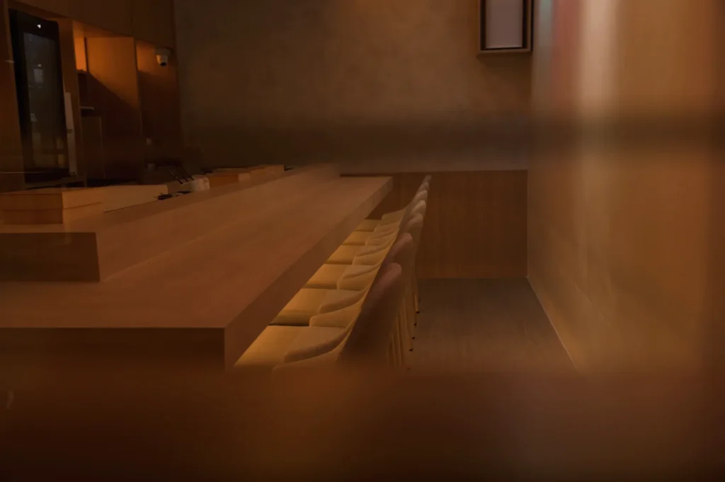 Dimly lit, cozy interior with a long wooden bar lined with plush chairs. Warm tones create an inviting, intimate atmosphere. Minimalist design.