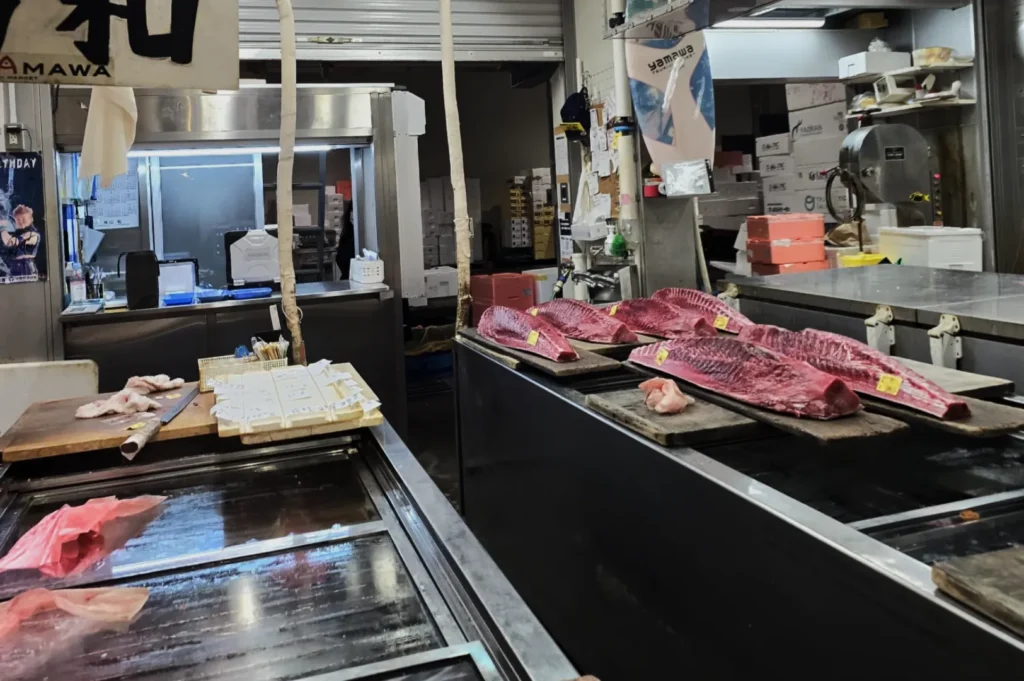 Indoor market scene showing large tuna cuts on wooden slabs at a fish stall. Surrounding the pieces are various seafood items and market tools, creating a bustling, authentic atmosphere.