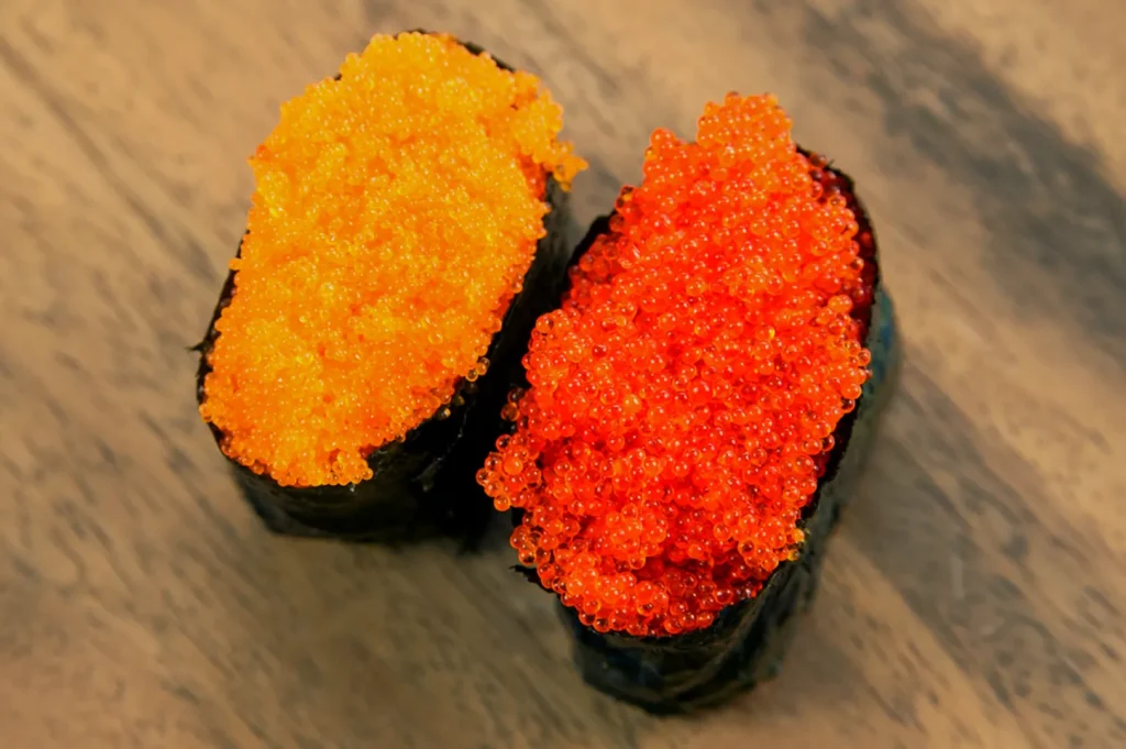 Two pieces of sushi topped with bright orange and red fish roe, wrapped in seaweed. The vibrant colors contrast with a light wooden background.