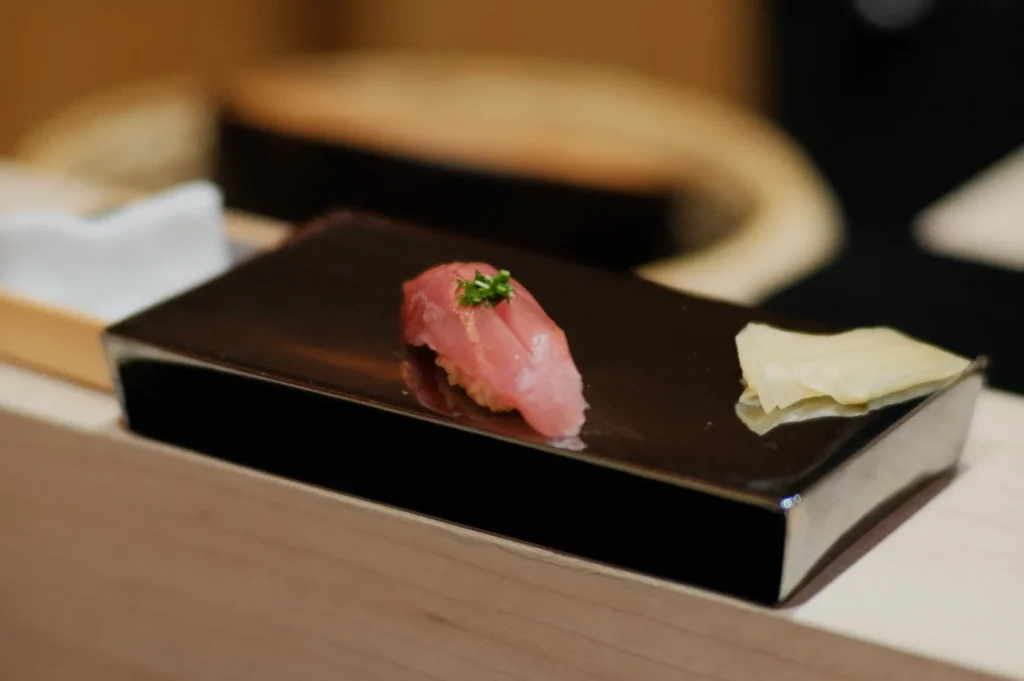 A piece of sushi with pink fish, garnished with herbs, is elegantly displayed on a sleek black plate alongside a slice of ginger. The setting conveys sophistication.