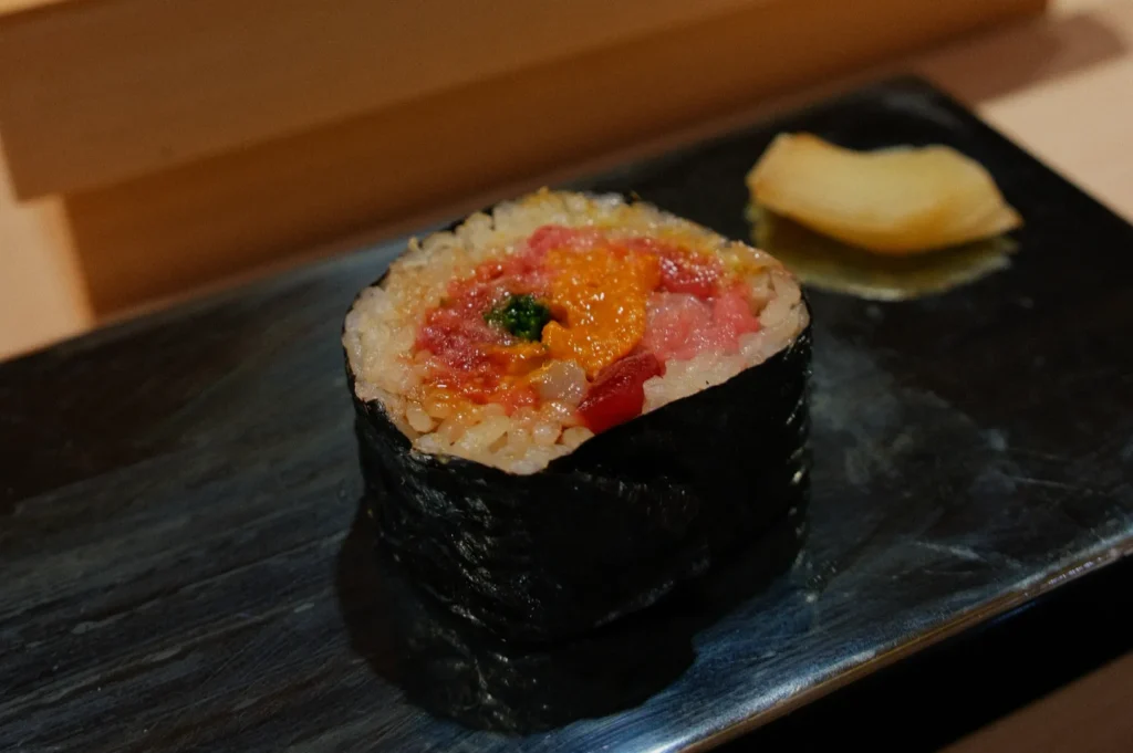 Single sushi roll on black plate, wrapped in seaweed, filled with rice and colorful toppings. Side of pickled ginger. Elegant, minimalist presentation.