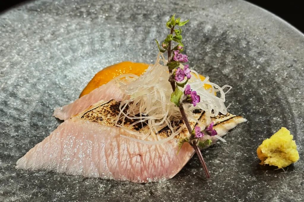 A delicate sashimi slice topped with shredded daikon and purple flowers is elegantly presented on a textured gray plate, conveying a refined, artistic vibe.