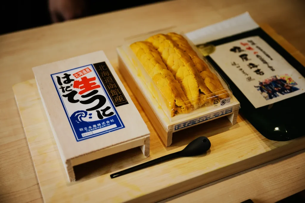 A neatly arranged wooden board displays a box of sea urchin (uni) in yellow hues, accompanied by a small black spoon and a package of condiments in Japanese packaging.