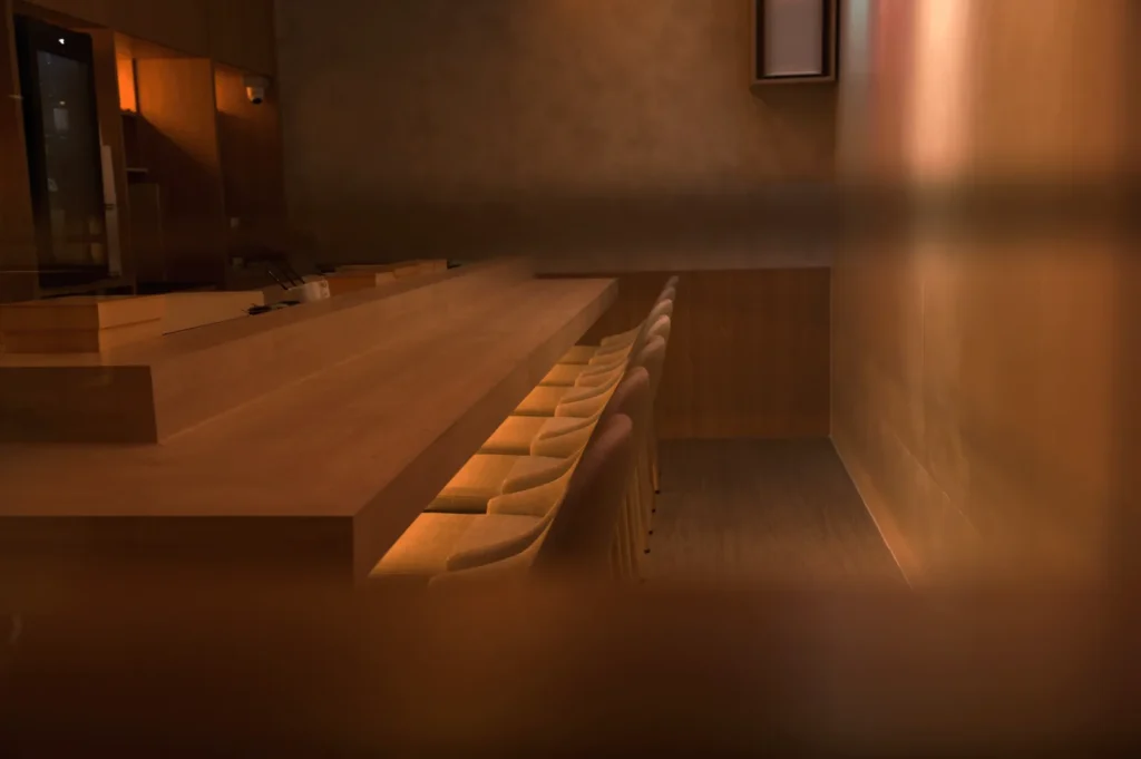 A dimly lit, cozy bar interior with a long wooden counter, neatly aligned chairs, and soft, warm lighting creating an intimate, inviting atmosphere.