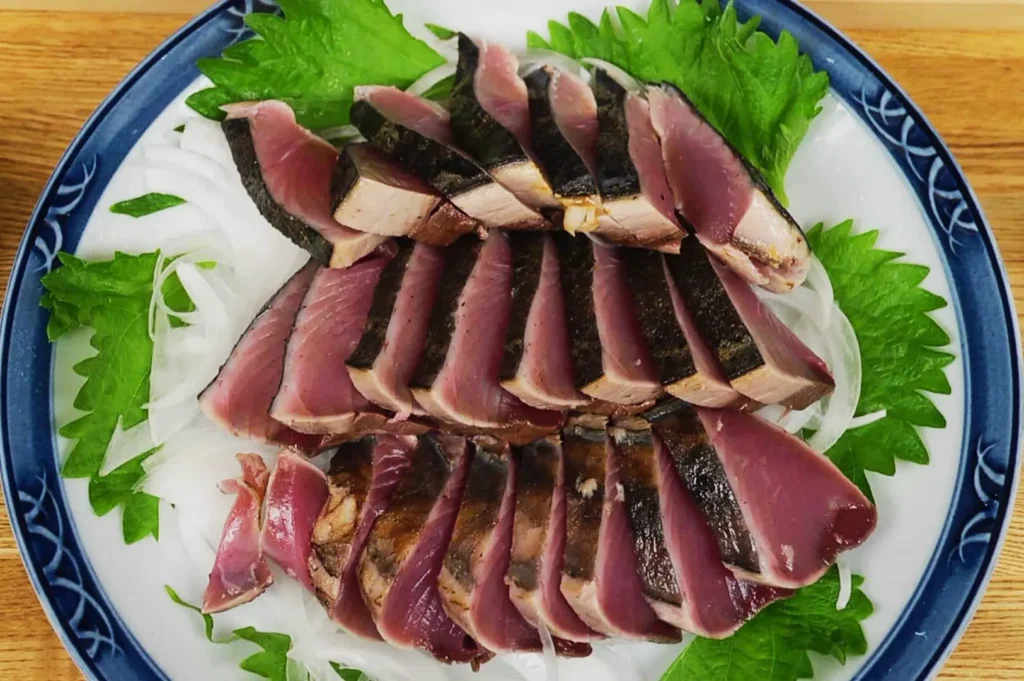 Sliced seared tuna arranged beautifully on a decorative blue and white plate, garnished with green shiso leaves and thinly sliced white onions.