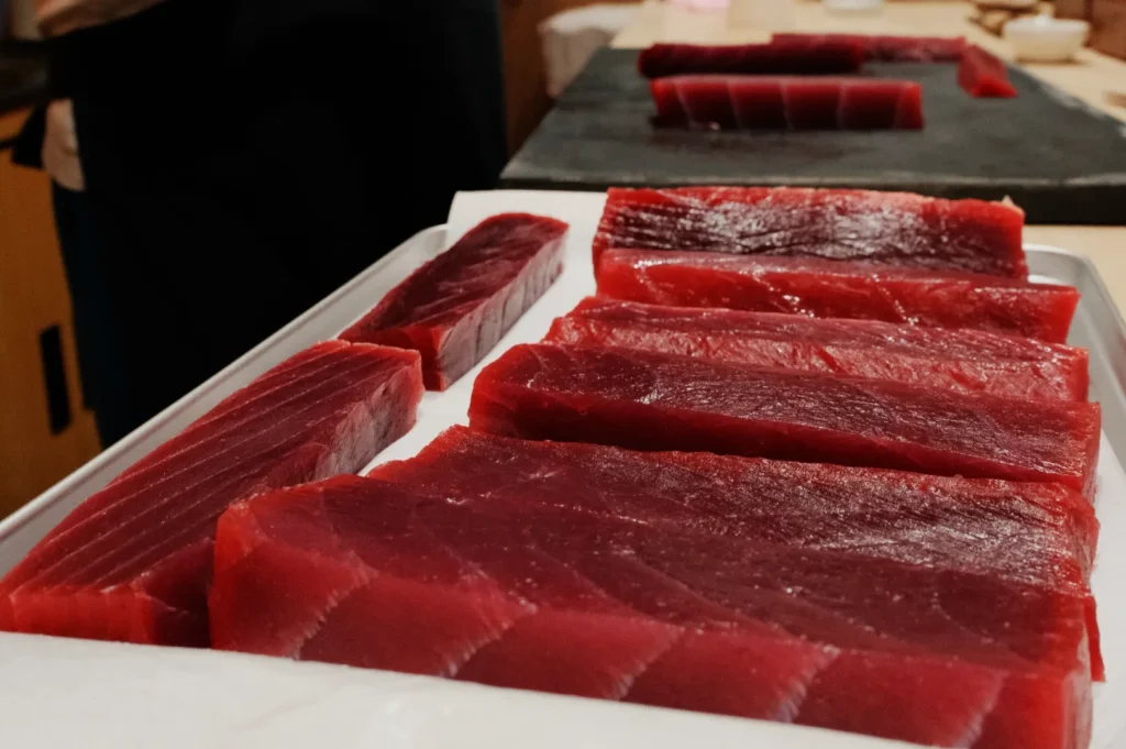 Close-up of neatly arranged slices of raw red tuna on a tray, displayed on a countertop. The scene conveys freshness and culinary precision.