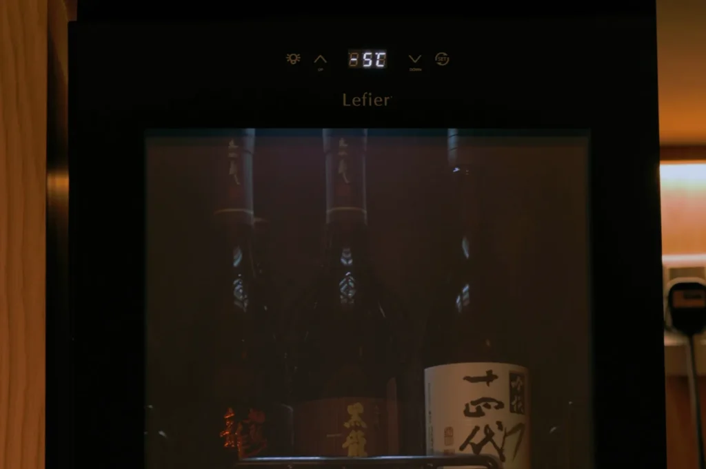 A dimly lit wine cooler displays three dark bottles inside. The digital temperature reading on top shows 5°C, creating a calm and sophisticated ambiance.