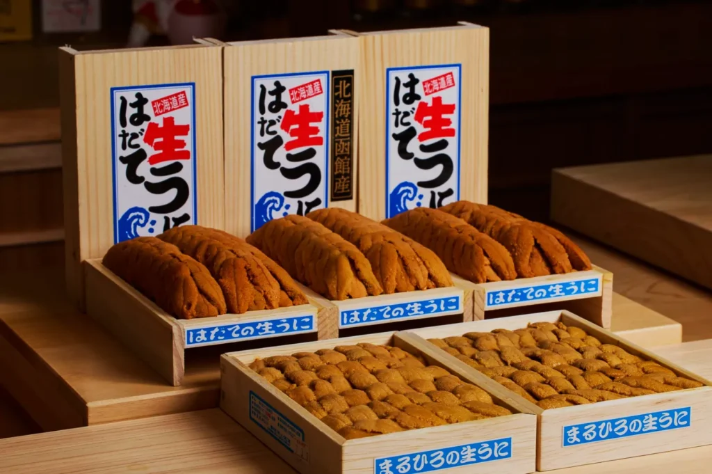 Wooden boxes of fresh sea urchin (uni) are displayed, with neat rows of orange uni. Japanese text and blue labels adorn the boxes. The scene conveys a sense of freshness and delicacy.