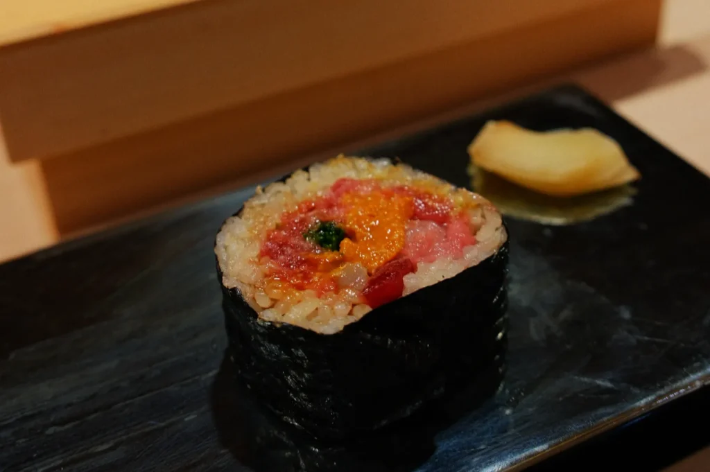 A close-up of a sushi roll placed on a dark plate, featuring vibrant orange, red, and pink fillings, with a side of pickled ginger. The scene evokes a sense of elegant dining.