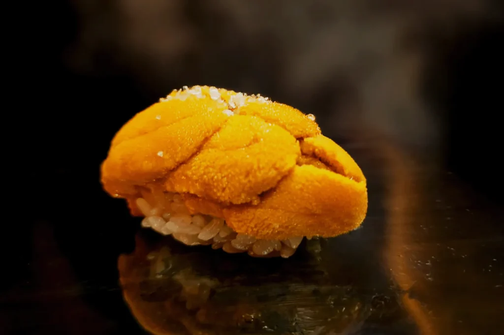 Close-up of a piece of uni sushi with vibrant orange sea urchin on a bed of white rice. The background is dark, highlighting the sushi's texture.