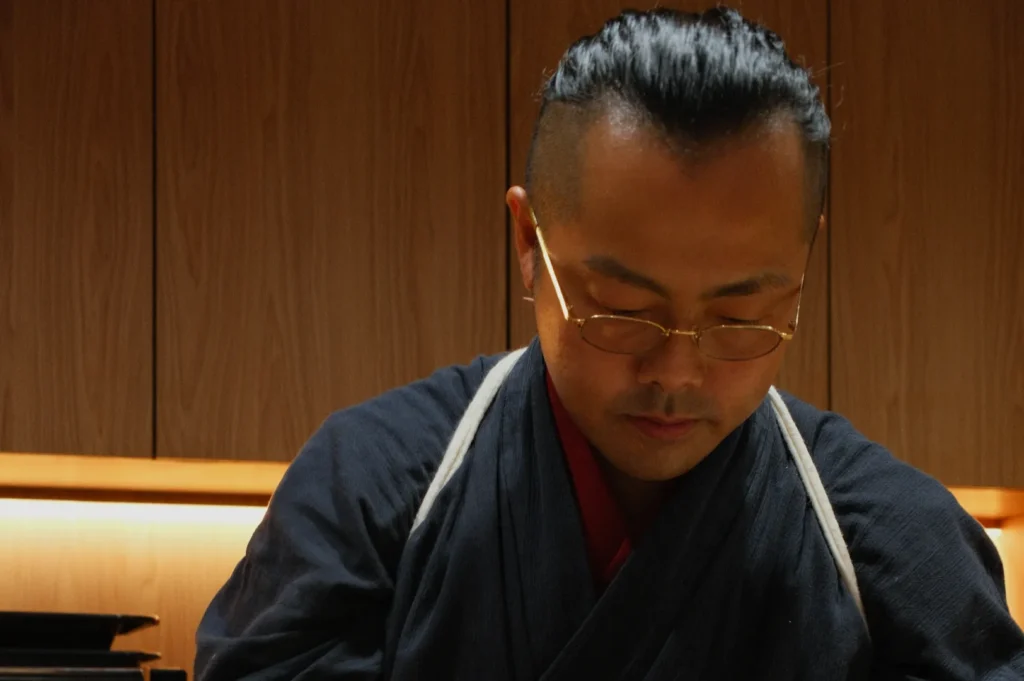 Chef Masa in traditional Japanese attire and glasses is focused, with a serene expression. They're in a warmly lit wooden room, exuding calmness.