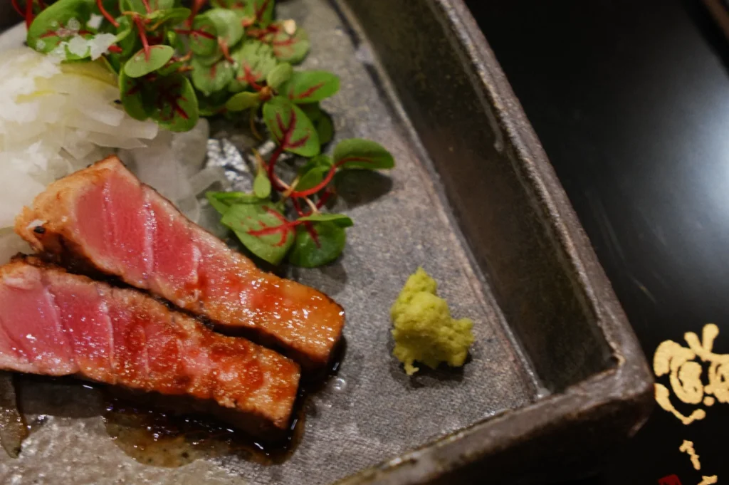Seared tuna slices on a gray plate, garnished with wasabi, radish greens, and shredded daikon. The dish has a fresh and elegant presentation.