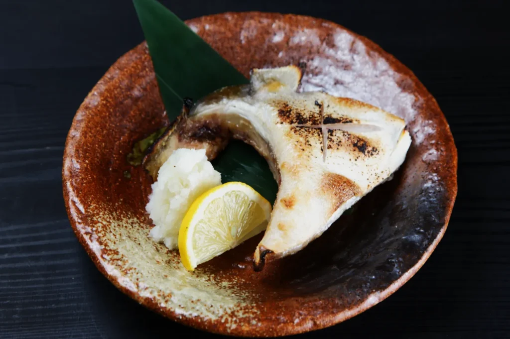 Grilled fish collar on a brown plate with a slice of lemon and grated daikon, garnished with a green leaf, set on a dark wooden table.