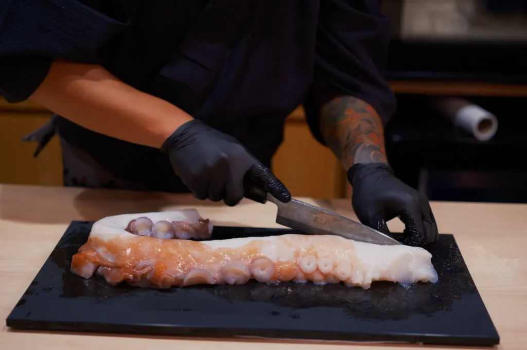 A chef wearing black gloves slices a large octopus tentacle on a black cutting board, showcasing precise culinary skill in a kitchen setting.