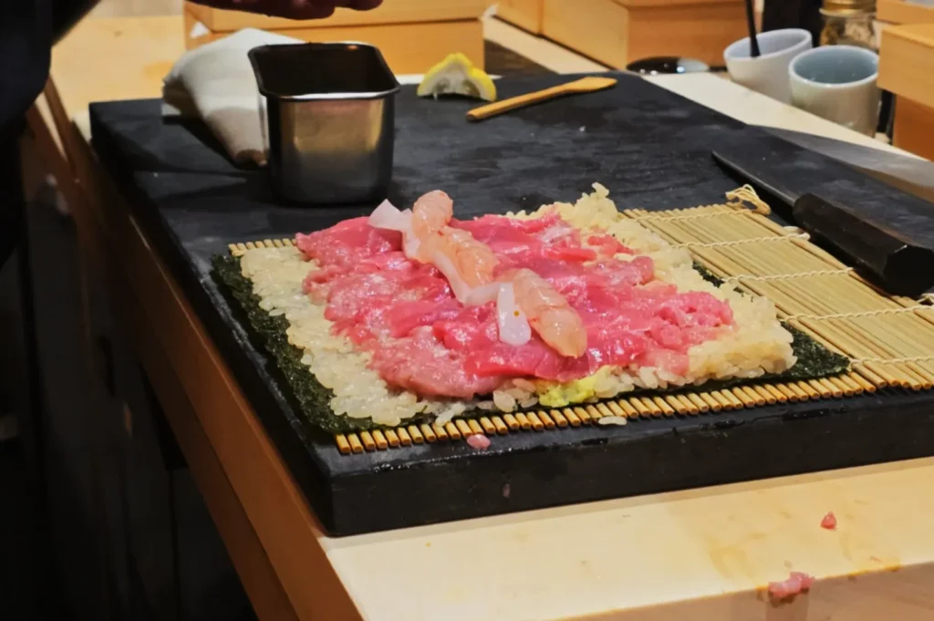 Sushi preparation on a bamboo mat. Rice and raw pink fish are layered on seaweed. A knife and metal cup are nearby, suggesting a sushi-making setting.