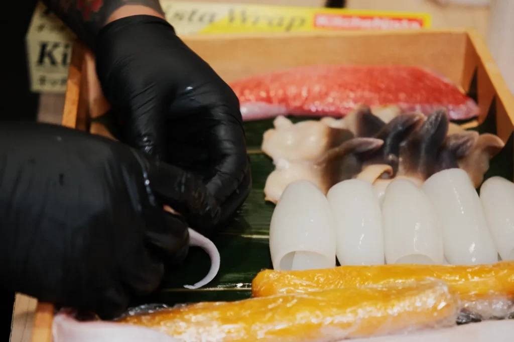 Hands in black gloves are arranging fresh seafood on a tray, featuring squid, fish, and shellfish, highlighting precision and care in preparation.