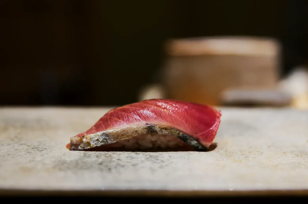 A piece of nigiri sushi featuring vivid pink fish over rice sits on a stone plate. The soft lighting and neutral background create a serene atmosphere.