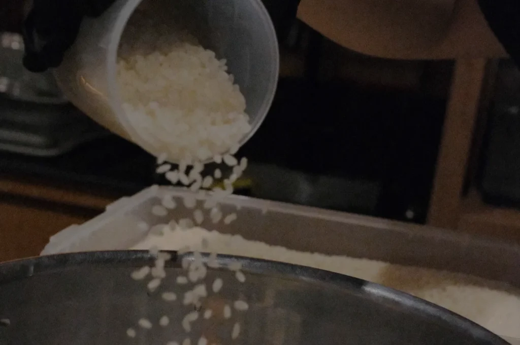 A hand in a dark glove pours uncooked rice from a cup into a metal bowl. The scene is dimly lit, conveying a focus on the action of cooking preparation.