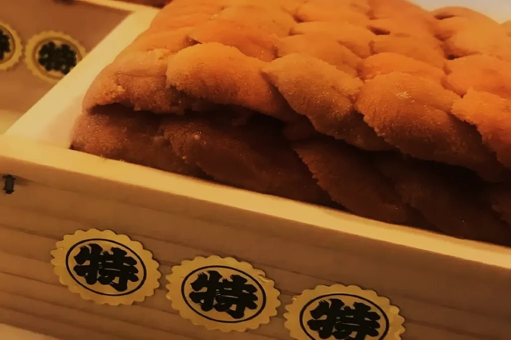 A wooden box containing neatly arranged, bright orange sea urchin roe (uni) with a rich, creamy texture, labeled with Japanese stickers.