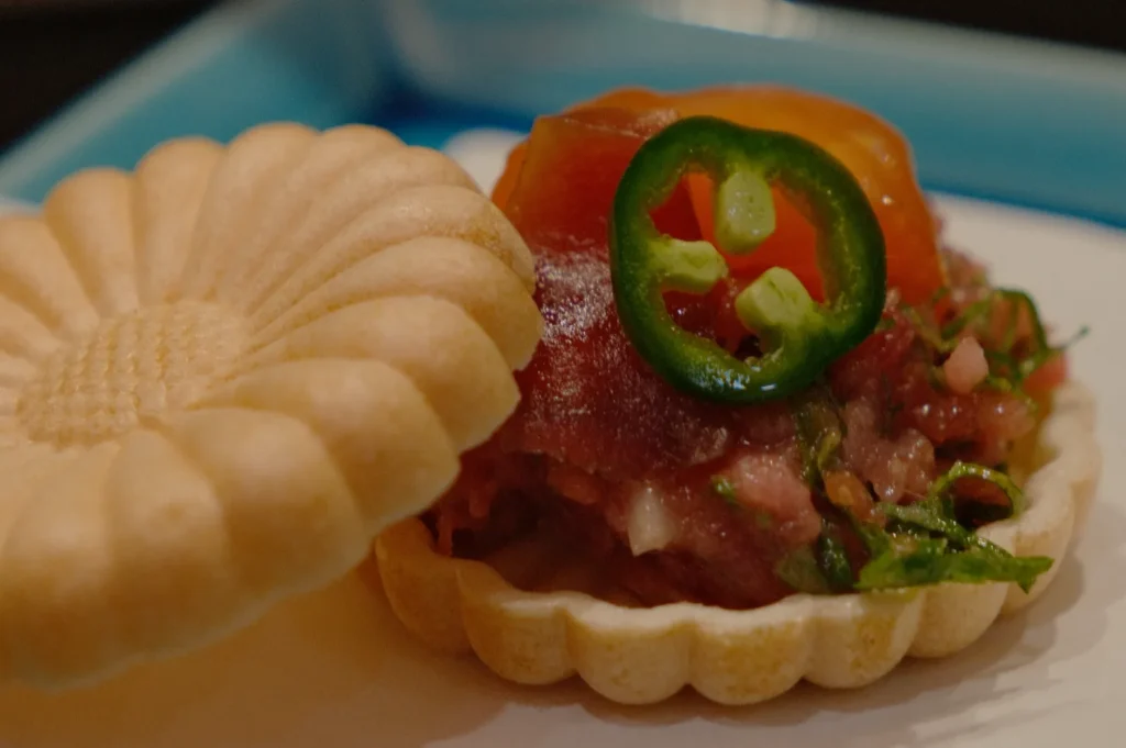 A gourmet dish featuring a flower-shaped wafer filled with raw tuna, topped with a jalapeño slice, tomato, and greens on a blue platter.