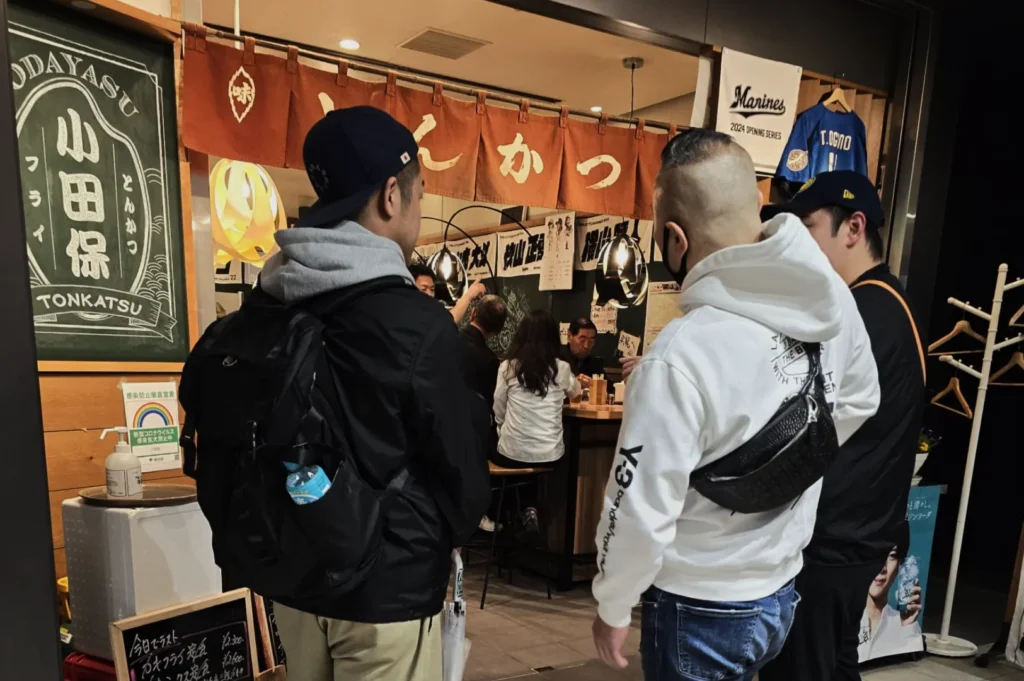 Three people are standing outside a cozy Japanese restaurant with wooden decor and a lively atmosphere. Inside, diners are seated at a counter.