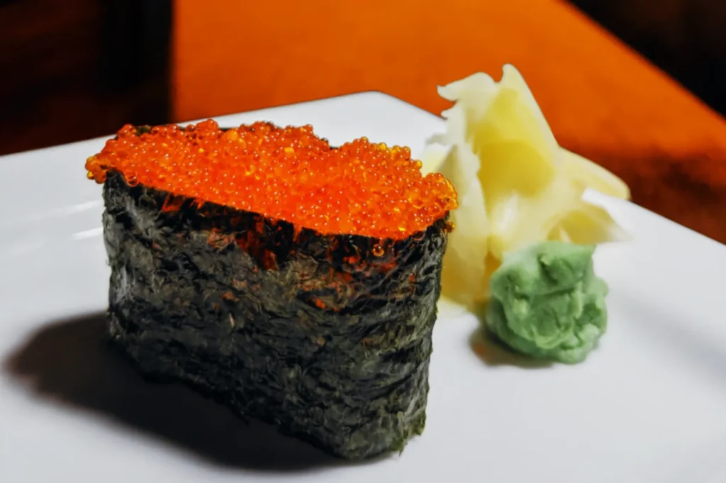 Gunkan maki sushi topped with bright orange fish roe on a white plate, accompanied by wasabi and pickled ginger. The setting is elegant and inviting.
