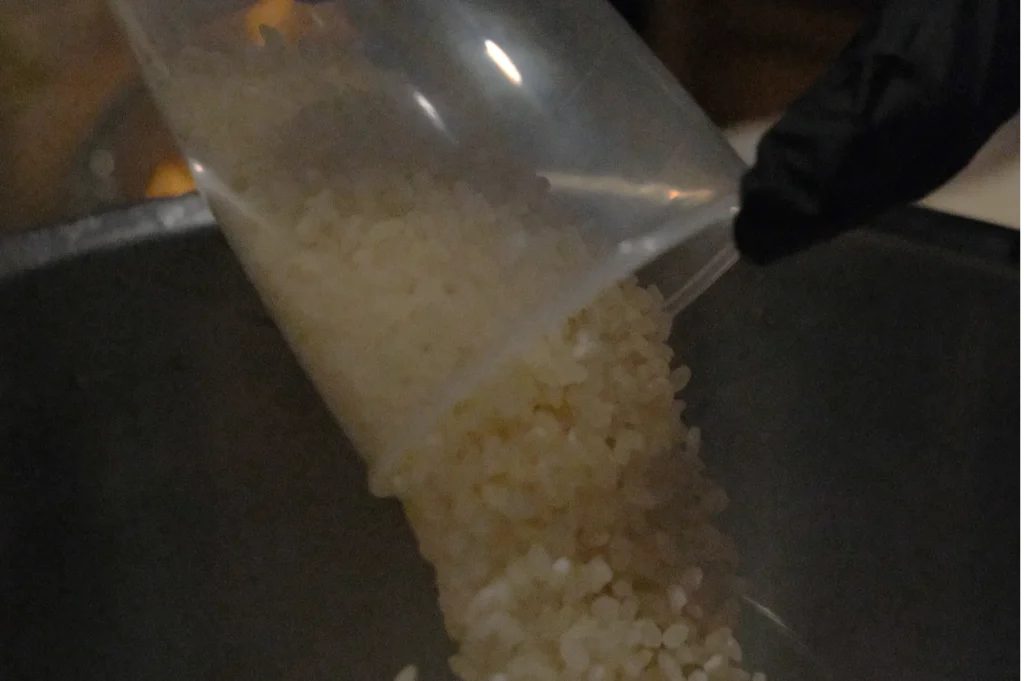 A person with a gloved hand pours uncooked rice from a clear plastic cup into a pot, captured in dim lighting, creating a focused, functional mood.
