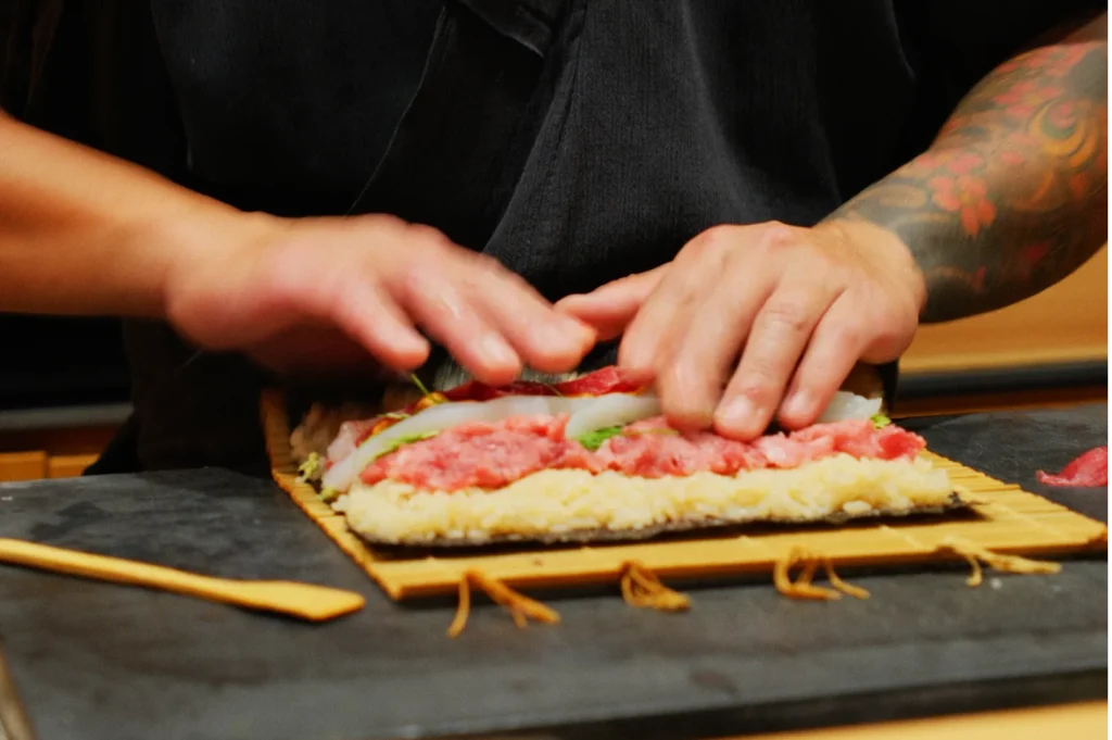 A person with tattooed arms skillfully rolls sushi on a bamboo mat, layering rice, fish, and vegetables. The scene conveys focus and culinary expertise.