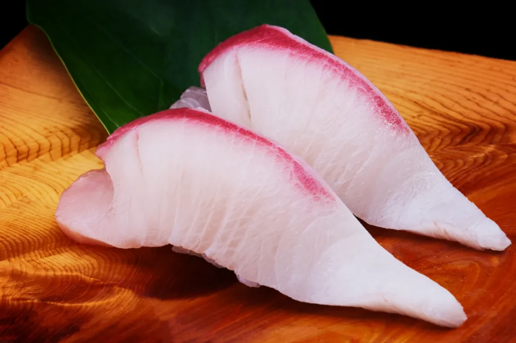 Two pieces of hamachi nigiri sushi on a wooden plate, with a vibrant green leaf as garnish. The fresh fish slices are creamy white with a pink hue.
