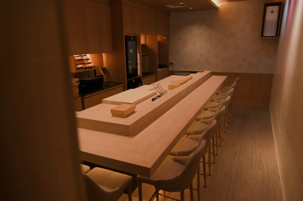 Elegant sushi bar with a long, wooden counter surrounded by chairs. Minimalist design with a warm, ambient glow and a neatly organized kitchen in the background.