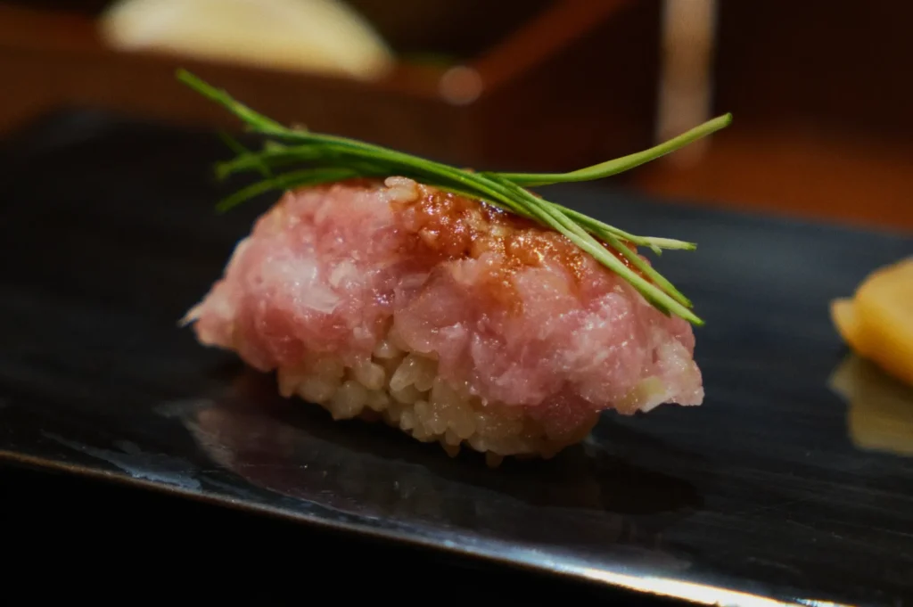 Close-up of a piece of sushi on a black plate, featuring pink minced fish topped with green chives, evoking freshness and elegance.