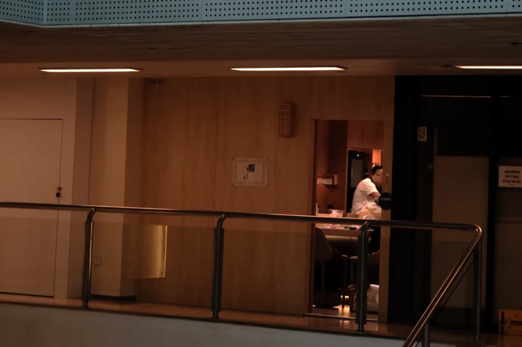Chef Masa in a white shirt works alone in a dimly lit room, viewed from a corridor. The atmosphere feels quiet and introspective, with warm lighting.