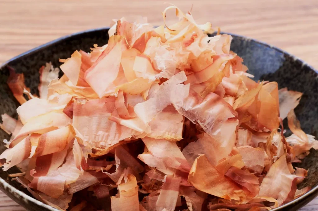 A dark bowl filled with delicate, thin pink and beige bonito flakes sits on a wooden table, conveying a light and airy texture.