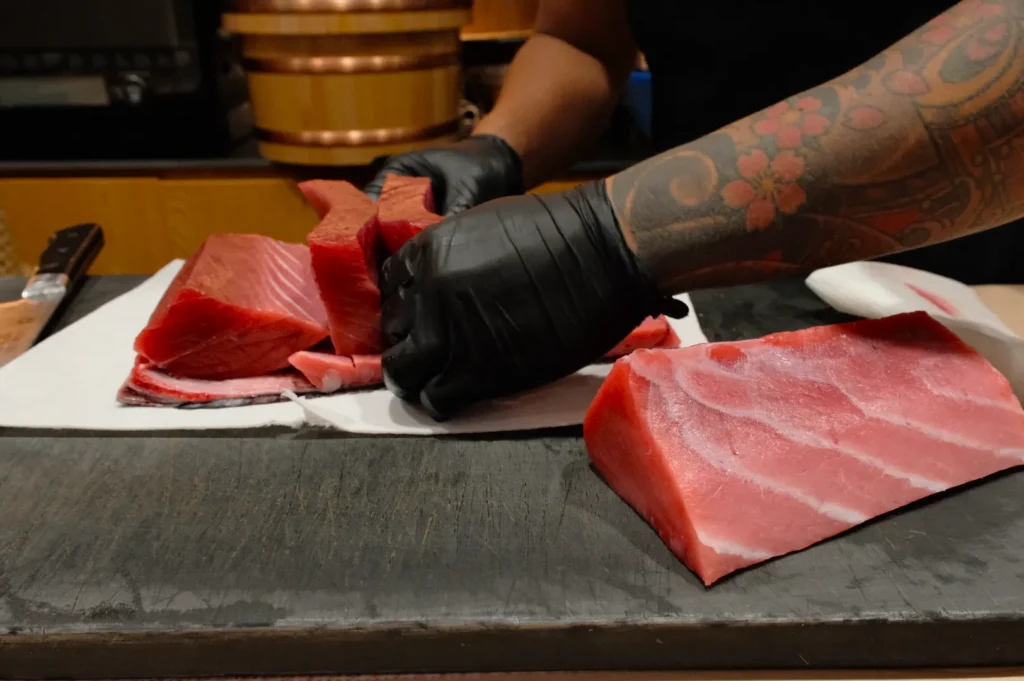 Person with tattooed arm and black gloves is slicing raw, red tuna on a chopping board. The setting conveys a focus on precise culinary preparation.