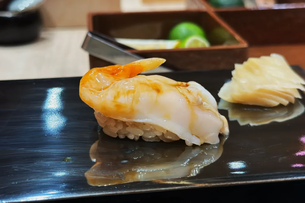 A piece of sushi with glossy, translucent seafood on rice, served on a reflective black plate with pickled ginger. Background includes limes in a box.