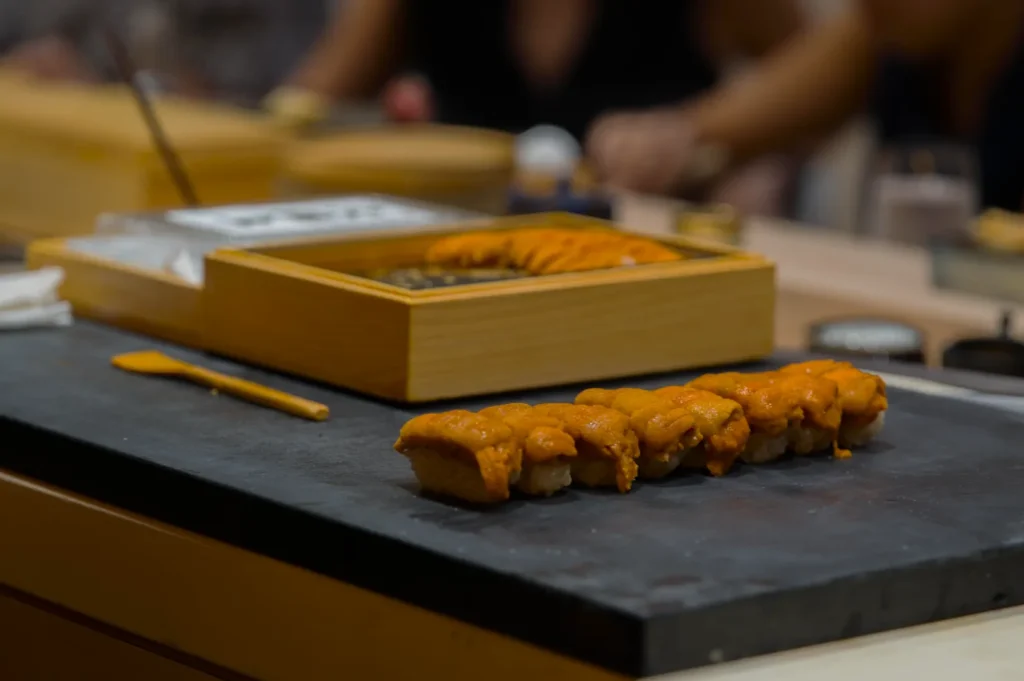 Close-up of uni sushi pieces on a black slate board with a wooden box of sea urchin in the background. The setting is elegant and minimalistic.