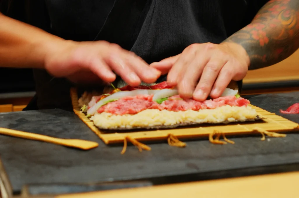 Hands with tattooed arms carefully roll sushi using a bamboo mat. The sushi features layers of rice, red fish, and vegetables, conveying focus and craftsmanship.