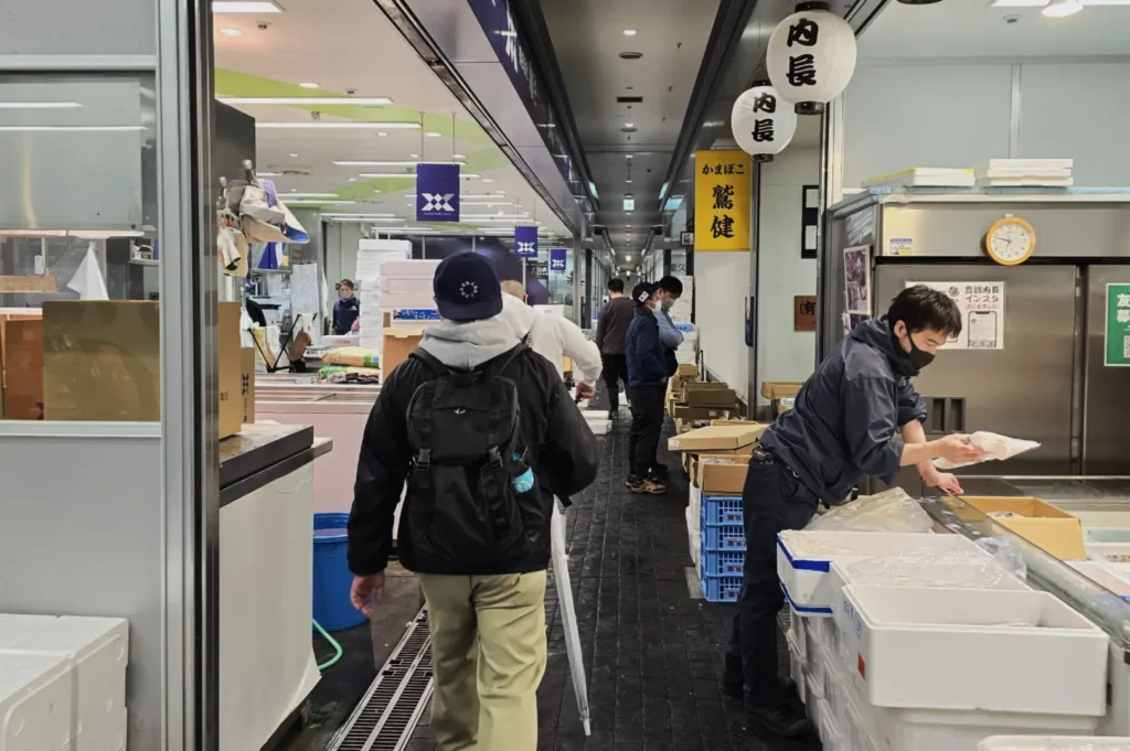 A bustling indoor market scene with people moving through aisles. Workers organize goods on both sides, creating a lively, busy atmosphere.