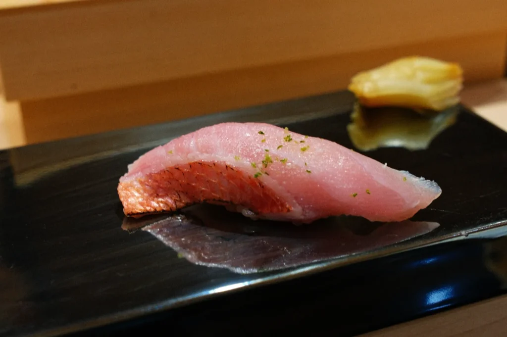 Close-up of a piece of sushi on a glossy black plate. The sushi features a slice of raw fish with delicate green seasoning, complemented by a small side of pickled ginger.