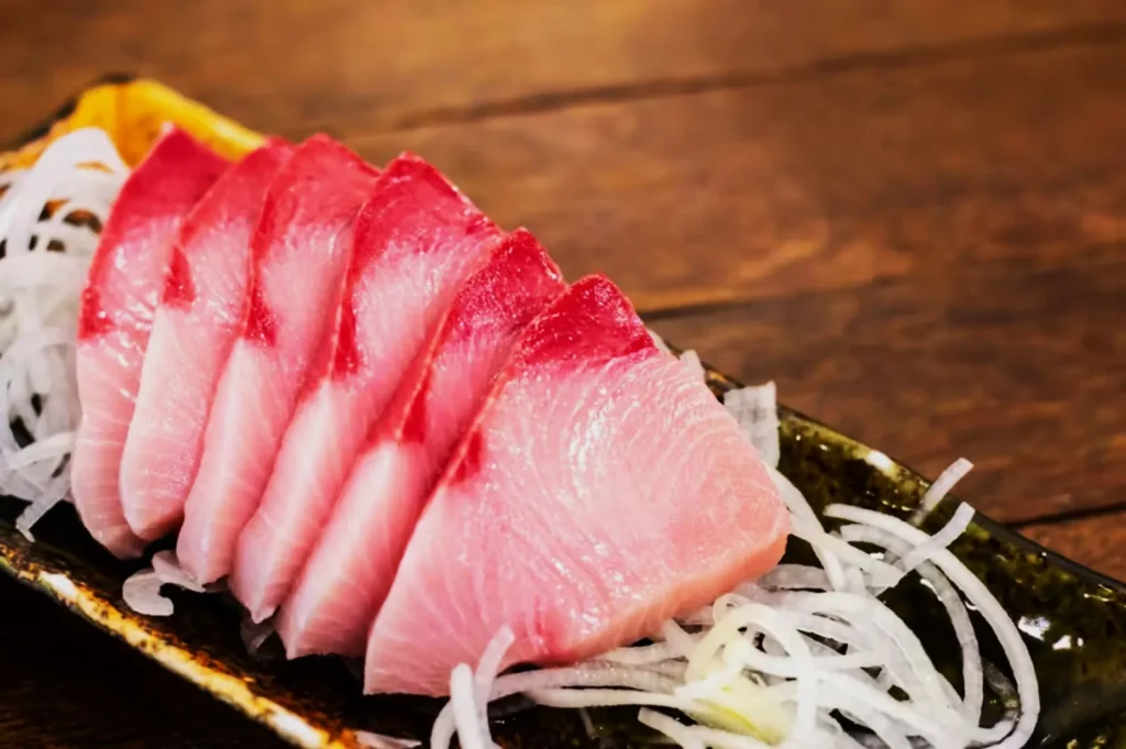 Slices of pink sashimi arranged on a dark rectangular plate with white shredded daikon radish. The setting is a wooden tabletop, evoking a fresh, appetizing feel.
