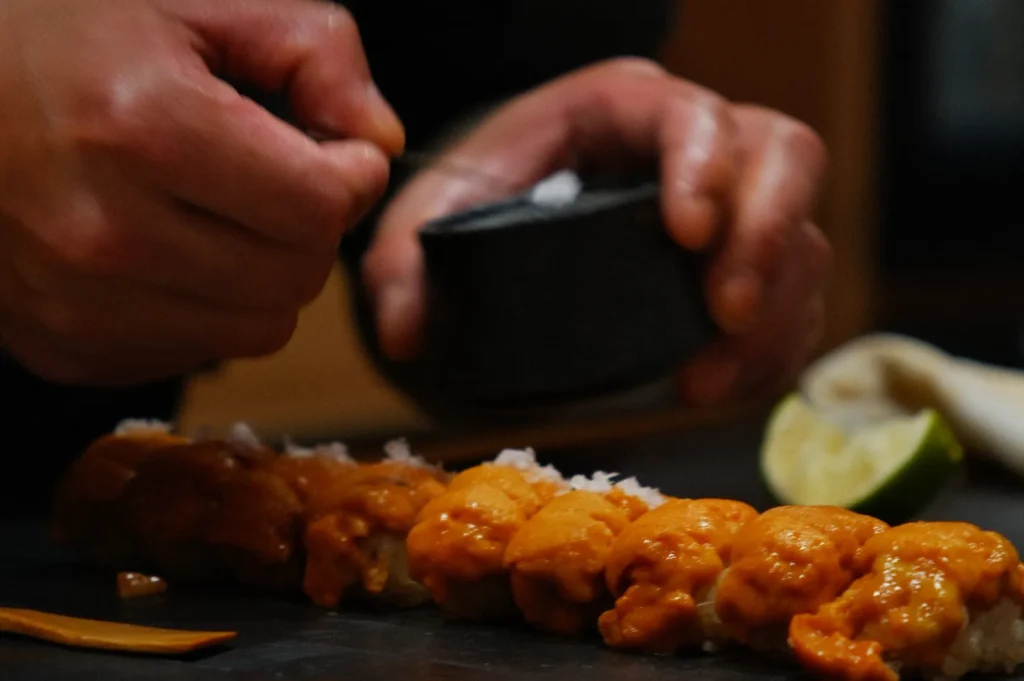 A chef's hands artfully sprinkle coarse salt onto a row of uni sushi. A halved lime is blurred in the background, adding a touch of freshness.
