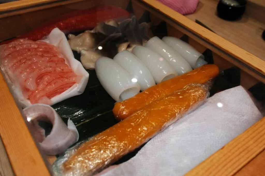 A wooden box contains an assortment of seafood, including translucent shrimp, white squid, yellow roe, and shellfish on green leaves, covered by a glass lid.