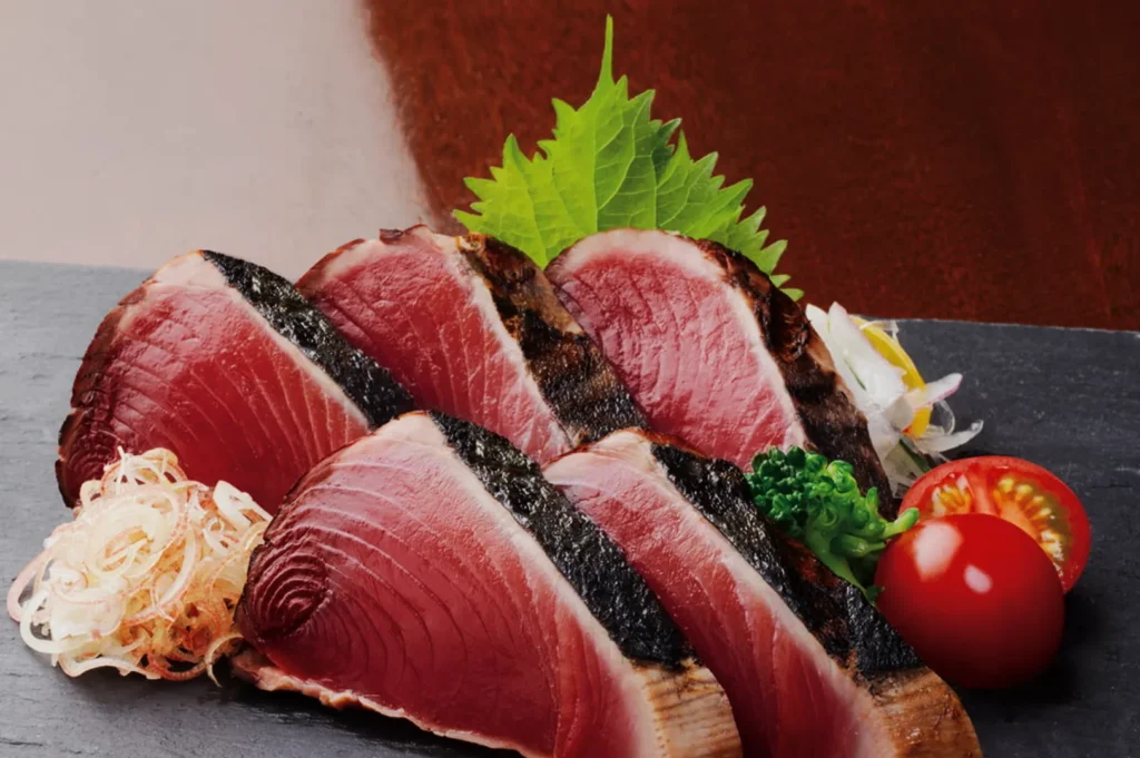 Slices of seared tuna sashimi arranged on a dark slate platter, garnished with a shiso leaf, shredded onions, and cherry tomatoes. Fresh and appetizing presentation.