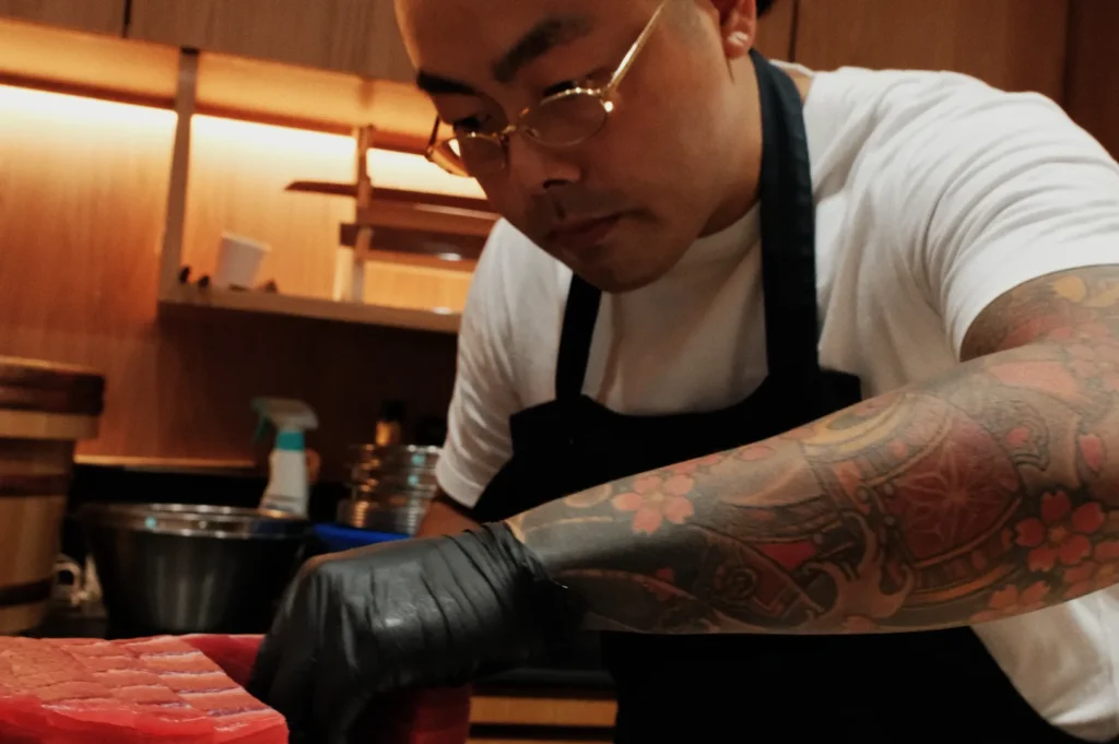 A chef with glasses and a tattooed arm concentrates as he slices raw tuna in a kitchen, conveying focus and artistry. Kitchen utensils are visible behind him.