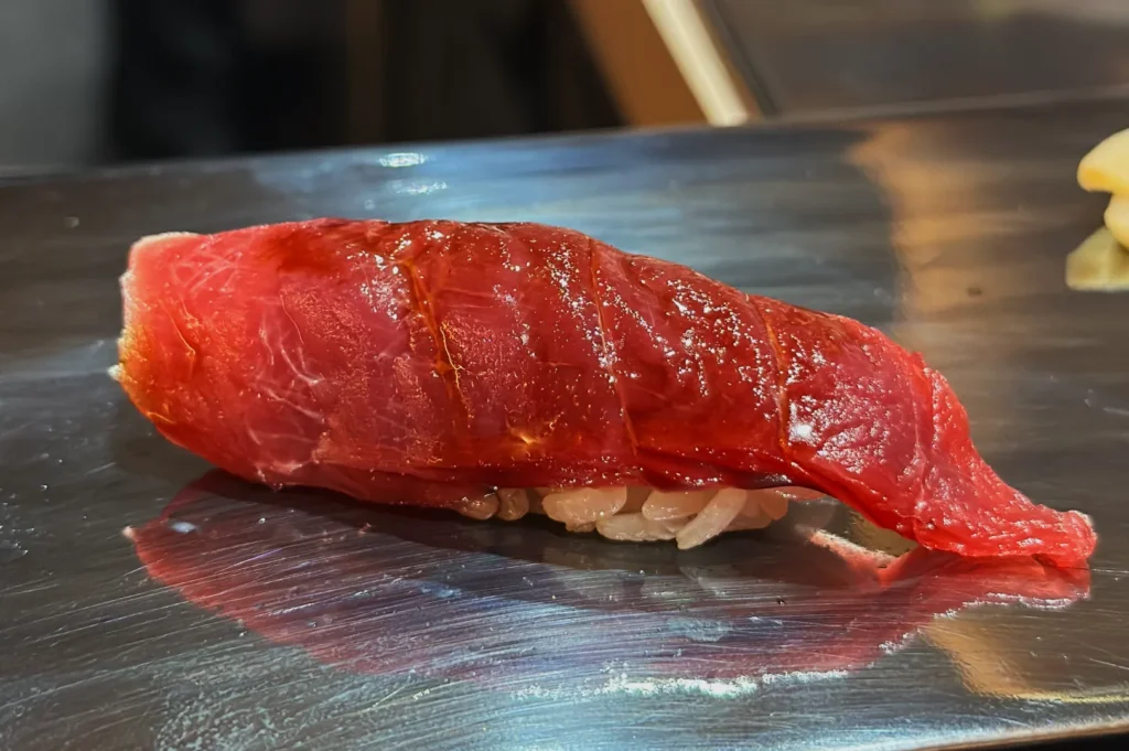 Close-up of a piece of red tuna sashimi on a bed of white rice, placed on a shiny metal surface. The fish looks fresh and vibrant.