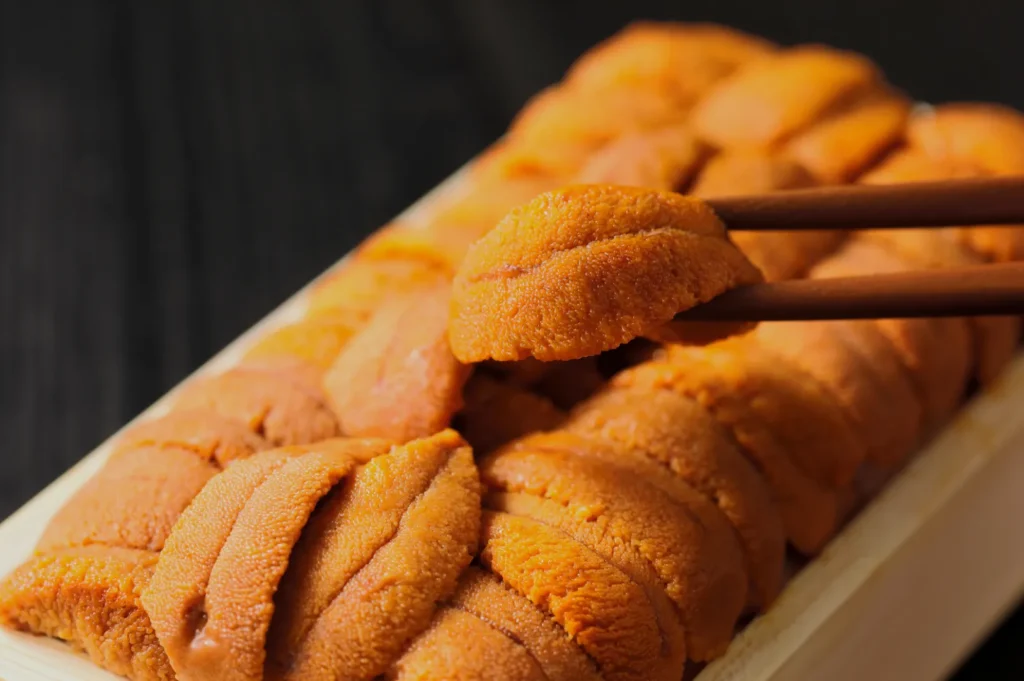 Close-up of fresh sea urchin roe (uni) in a wooden tray, with chopsticks holding one piece. The rich orange color contrasts with the dark background.