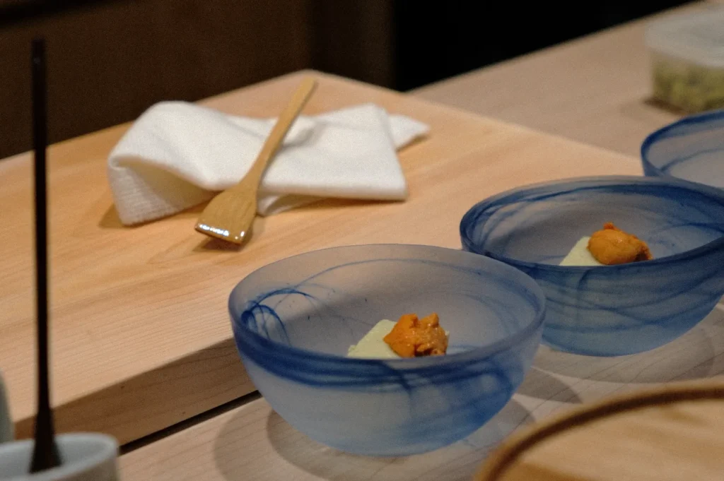 Blue bowls on a wooden table each hold a small portion of orange uni and green garnish. A folded napkin with a wooden spoon sits nearby. Elegant and minimal.