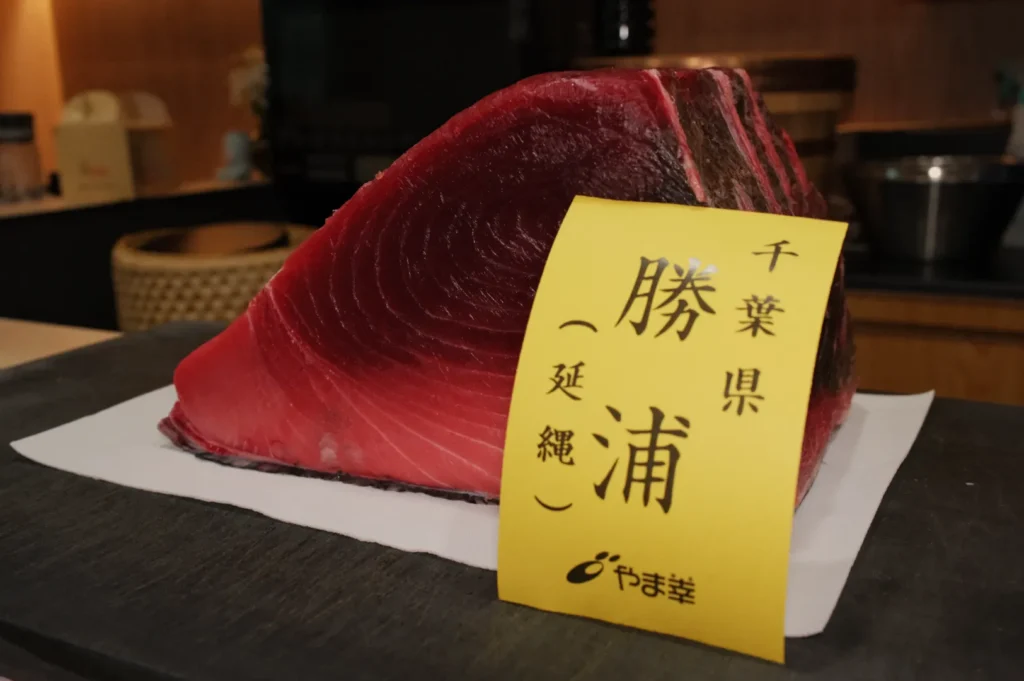 A thick, marbled slice of fresh tuna is displayed on a white surface, with a yellow Japanese label attached. The setting conveys a sense of culinary expertise.