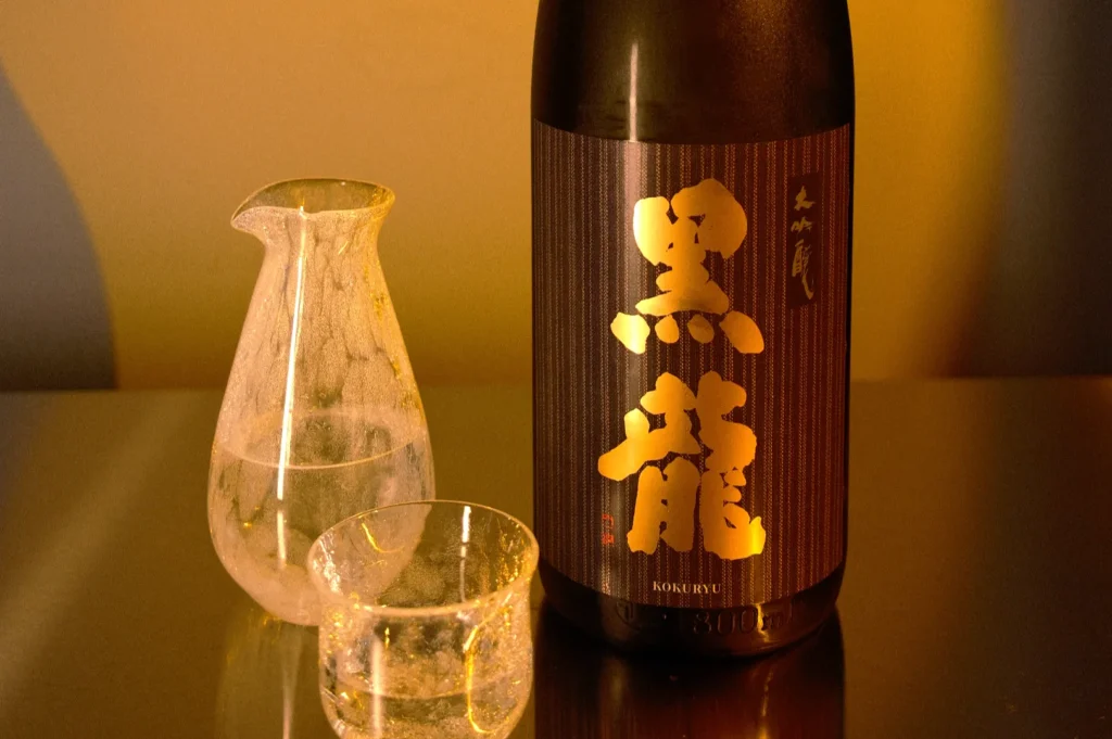 A bottle of sake with bold Japanese characters sits on a reflective surface next to a clear carafe and glass, set in warm lighting, evoking a cozy atmosphere.